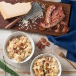 two bowls of bacon mac and cheese sit ready to eat next to a wooden cutting board that holds grated white cheese and cooked bacon. A gold for sits nearby,