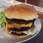 cheeseburger stacked with three patties and two types of cheese on a white plate with caesar salad
