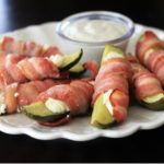 eight pickle spears wrapped in cooked bacon sitting on a white plate with a small bowl of dip
